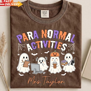 May include: A brown t-shirt featuring the text "PARA NORMAL ACTIVITIES" in purple, orange, and white. The graphic includes four ghosts, one with a pumpkin on its head, and the name "Mrs Taylor" in orange script. The shirt has a Comfort Colors label.
