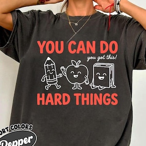 May include: A charcoal gray short-sleeve t-shirt featuring the orange text YOU CAN DO HARD THINGS and the white text you got this! with cartoon illustrations of a smiling pencil, an apple, and a book. The shirt has a relaxed, loose fit.