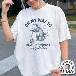 May include: White t-shirt with a graphic of an opossum riding a scooter, with the text "ON MY WAY TO HELP TINY HUMANS regulate". The design is in shades of blue. The shirt has a Comfort Colors label.