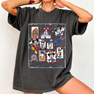 May include: A charcoal gray short-sleeve t-shirt featuring a large rectangular graphic print. The design includes various Disney characters like Mickey, Minnie, Donald, and Goofy in patriotic red, white, and blue outfits, alongside a castle and the text 2026.