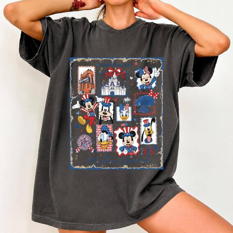 May include: A charcoal gray short-sleeve t-shirt featuring a large rectangular graphic print. The design includes various Disney characters like Mickey, Minnie, Donald, and Goofy in patriotic red, white, and blue outfits, alongside a castle and the text 2026.