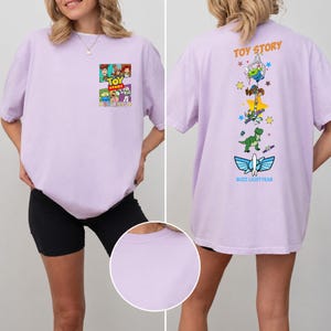 May include: A light purple t-shirt featuring a Toy Story graphic on the front left chest with the text Toy Story Est 1995. The back displays a vertical design of Toy Story characters including Buzz Lightyear and Rex with the text Toy Story.