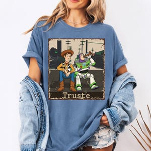 Peut inclure: Un t-shirt bleu avec une impression graphique repr&eacute;sentant les personnages Woody et Buzz l'&Eacute;clair assis ensemble. Le motif pr&eacute;sente une bordure vieillie et inclut le texte "Truste" en bas dans une police serif grasse.