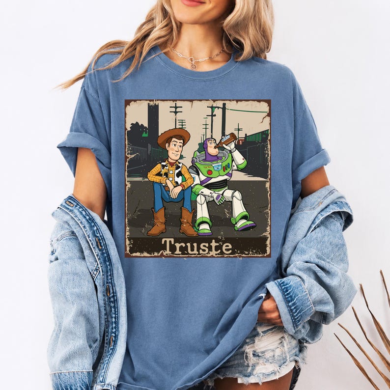 Peut inclure: Un t-shirt bleu avec une impression graphique repr&eacute;sentant les personnages Woody et Buzz l'&Eacute;clair assis ensemble. Le motif pr&eacute;sente une bordure vieillie et inclut le texte "Truste" en bas dans une police serif grasse.
