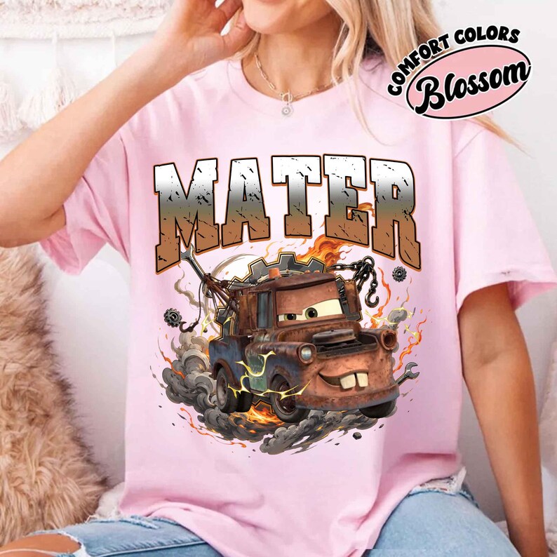 K&ouml;nnte beinhalten: Ein hellrosa T-Shirt mit einem Cartoon-Bild von Mater aus dem Film Cars. Das Wort "MATER" ist in einem Farbverlauf &uuml;ber dem Bild aufgedruckt. Auf dem Shirt stehen auch die Worte "COMFORT COLORS Blossom" rechts oben.