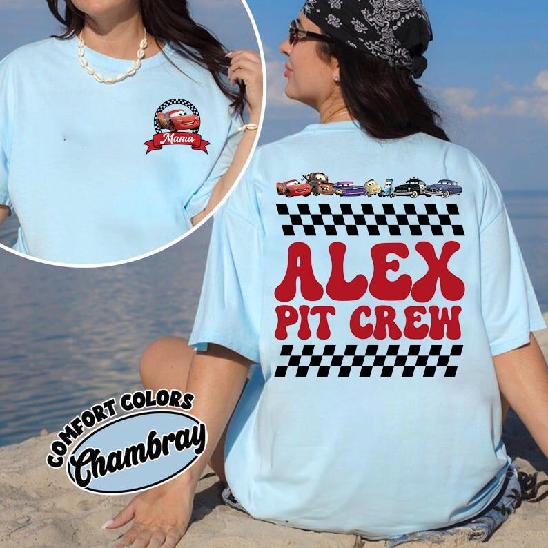 May include: A light blue t-shirt featuring a small graphic on the front chest of a red race car with the text Mama. The back displays a row of colorful race car characters above a checkered pattern, the red text ALEX PIT CREW, and another checkered pattern.