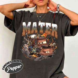 K&ouml;nnte beinhalten: Dunkelgraues T-Shirt mit einer Grafik von Hook aus dem Film Cars, mit dem Wort "MATER" &uuml;ber dem Bild. Das Design umfasst Flammen, Rauch und Blitzeffekte. Das Shirt tr&auml;gt die Aufschrift "COMFORT COLORS" und "Pepper".