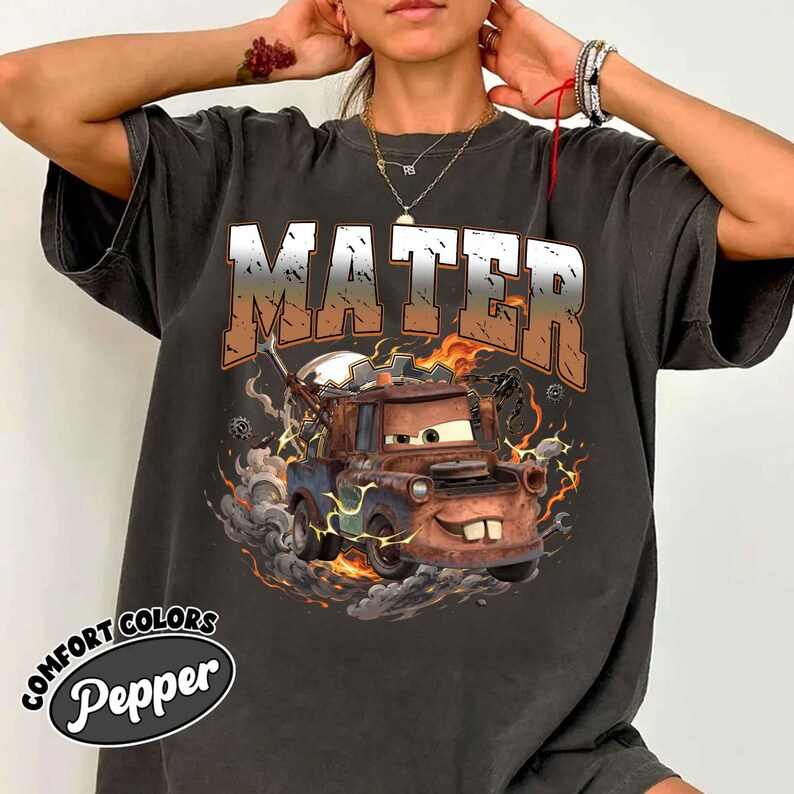 K&ouml;nnte beinhalten: Dunkelgraues T-Shirt mit einer Grafik von Hook aus dem Film Cars, mit dem Wort "MATER" &uuml;ber dem Bild. Das Design umfasst Flammen, Rauch und Blitzeffekte. Das Shirt tr&auml;gt die Aufschrift "COMFORT COLORS" und "Pepper".