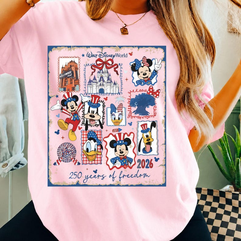 May include: A light pink t-shirt featuring a Walt Disney World graphic with various characters including Mickey Mouse, Minnie Mouse, Donald Duck, Daisy Duck, Goofy, and Pluto in patriotic attire, with the text 2026 and 250 years of freedom.