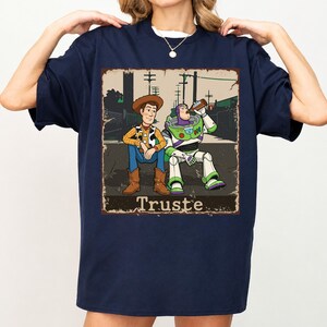Peut inclure: Un t-shirt bleu marine avec une impression graphique repr&eacute;sentant deux personnages de dessin anim&eacute; assis par terre. L'un porte un chapeau de cow-boy et un gilet, l'autre est un ranger de l'espace buvant &agrave; une bouteille. Le texte "Truste" est imprim&eacute; en dessous.
