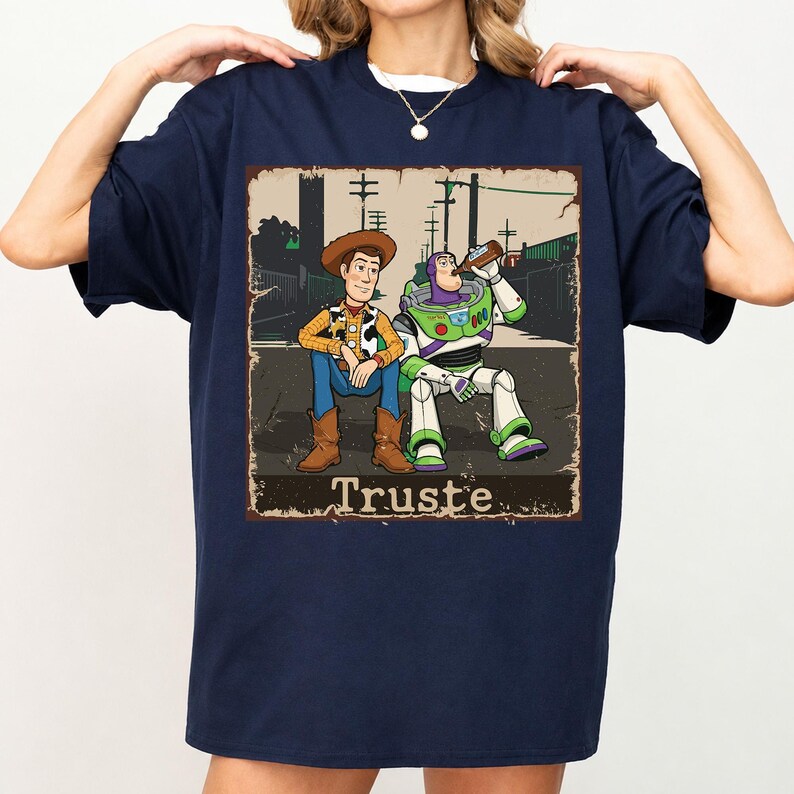 Peut inclure: Un t-shirt bleu marine avec une impression graphique repr&eacute;sentant deux personnages de dessin anim&eacute; assis par terre. L'un porte un chapeau de cow-boy et un gilet, l'autre est un ranger de l'espace buvant &agrave; une bouteille. Le texte "Truste" est imprim&eacute; en dessous.