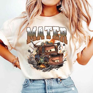 K&ouml;nnte beinhalten: Beiges T-Shirt mit einer Grafik von Hook aus dem Film Cars. Das Design zeigt das Wort "MATER" in einer verwitterten Schriftart, mit Flammen und Raucheffekten um den Zeichentrick-Truck.