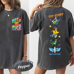 May include: A charcoal gray t-shirt featuring a small Toy Story graphic on the front chest with the text EST. 1995. The back displays a vertical design of Toy Story characters, including an alien, Woody, Buzz Lightyear, and Rex, with the text TOY STORY.