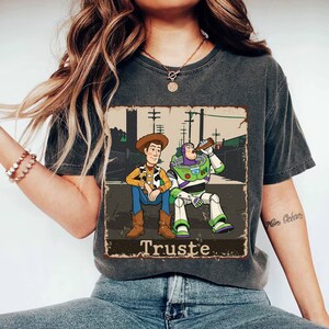 Peut inclure: Un t-shirt gris anthracite avec une impression graphique repr&eacute;sentant deux personnages de dessin anim&eacute; assis ensemble. L'un porte un chapeau de cow-boy et une chemise jaune, l'autre est un ranger de l'espace en costume blanc et vert. Le texte "Truste" est imprim&eacute; en dessous.
