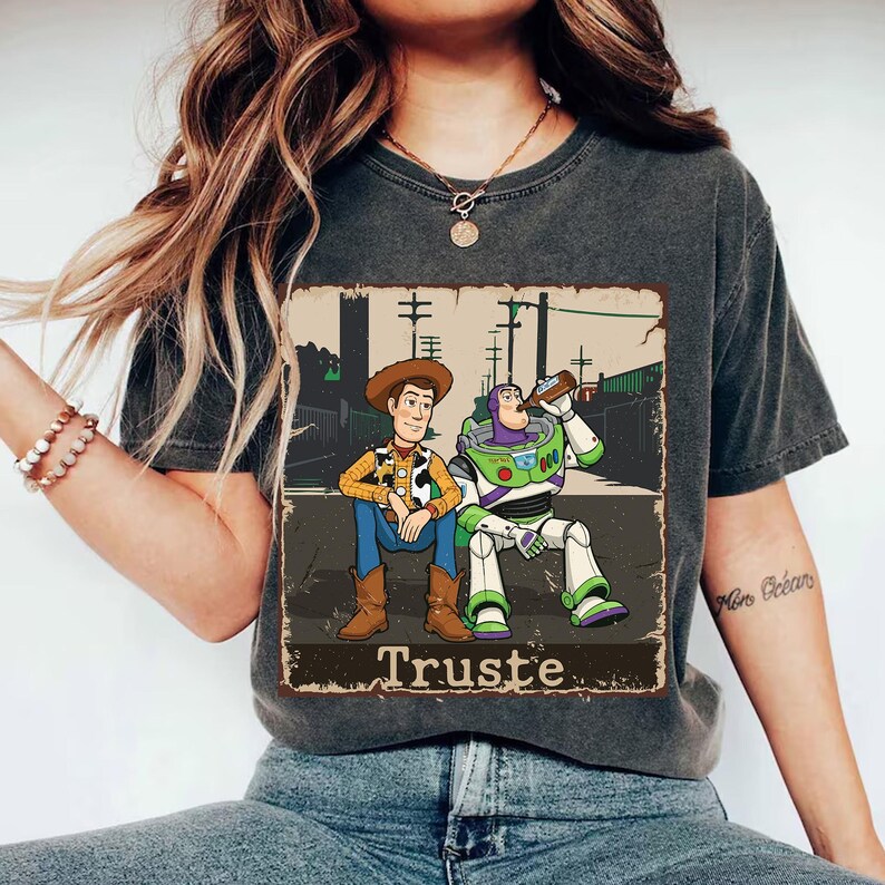 Peut inclure: Un t-shirt gris anthracite avec une impression graphique repr&eacute;sentant deux personnages de dessin anim&eacute; assis ensemble. L'un porte un chapeau de cow-boy et une chemise jaune, l'autre est un ranger de l'espace en costume blanc et vert. Le texte "Truste" est imprim&eacute; en dessous.