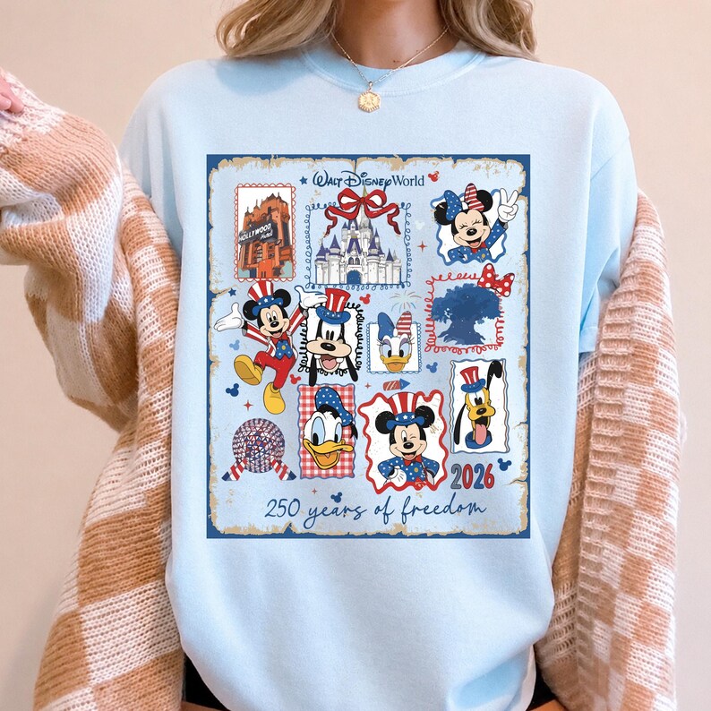 May include: A light blue sweatshirt featuring a graphic print with the text Walt Disney World and 250 years of freedom. The design includes illustrations of Mickey Mouse, Minnie Mouse, Donald Duck, Goofy, Pluto, and Daisy Duck in patriotic attire.