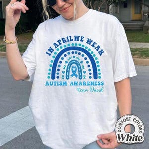 May include: White t-shirt with a blue and teal rainbow design. The shirt features the text "IN APRIL WE WEAR AUTISM AWARENESS #Team David". The design includes a ribbon and heart symbols.