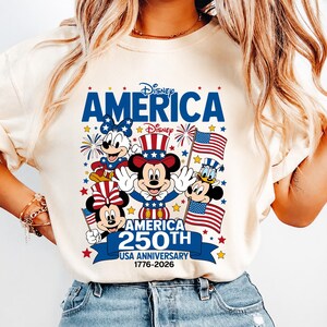 May include: A cream-coloured t-shirt featuring a graphic of Mickey Mouse, Minnie Mouse, and Donald Duck in patriotic attire. The text reads Disney AMERICA, Disney, AMERICA 250TH USA ANNIVERSARY 1776-2026, surrounded by stars, fireworks, and American flags.