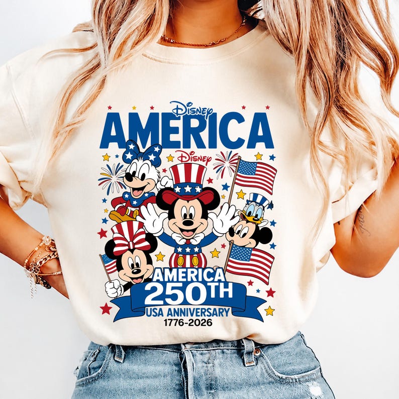 May include: A cream-coloured t-shirt featuring a graphic of Mickey Mouse, Minnie Mouse, and Donald Duck in patriotic attire. The text reads Disney AMERICA, Disney, AMERICA 250TH USA ANNIVERSARY 1776-2026, surrounded by stars, fireworks, and American flags.