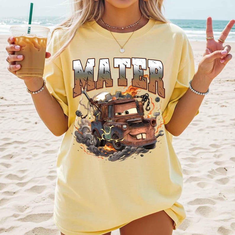 K&ouml;nnte beinhalten: Hellgelbes T-Shirt mit einer Grafik der Figur Mater aus dem Film Cars. Das Wort "MATER" ist &uuml;ber dem Bild des Abschleppwagens aufgedruckt. Das Design umfasst Flammen und Rauch, was dem T-Shirt ein dynamisches Aussehen verleiht.