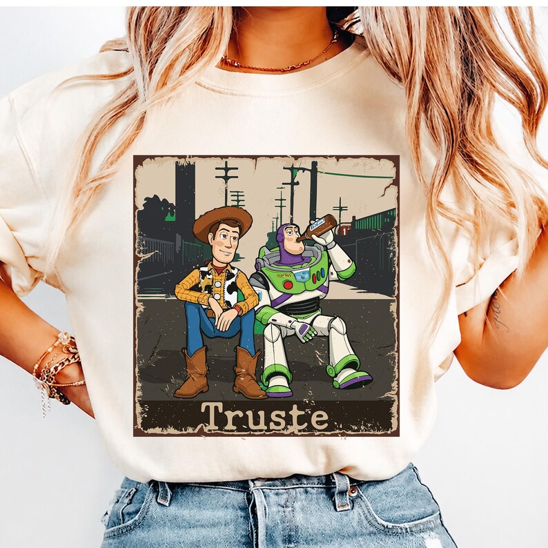 Peut inclure: Un t-shirt couleur cr&egrave;me avec une impression graphique repr&eacute;sentant deux personnages de dessin anim&eacute; assis ensemble. L'un porte un chapeau de cow-boy et un gilet, l'autre est un ranger de l'espace buvant &agrave; une bouteille. Le texte "Truste" est imprim&eacute;.
