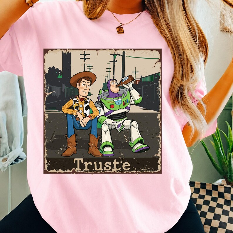 Peut inclure: Un t-shirt rose clair avec une impression graphique de Woody et Buzz l'&Eacute;clair assis ensemble. Woody porte un chapeau de cowboy, une chemise jaune et un jean bleu. Buzz porte une combinaison spatiale et boit dans une bouteille brune. Le texte "Truste" est en dessous.
