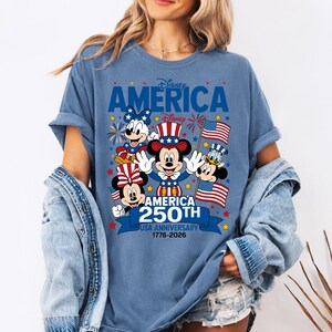 May include: A blue t-shirt featuring a graphic of Mickey Mouse, Minnie Mouse, and Donald Duck in patriotic attire. The design includes the text Disney AMERICA, Disney, AMERICA 250TH USA ANNIVERSARY 1776-2026, American flags, and fireworks.