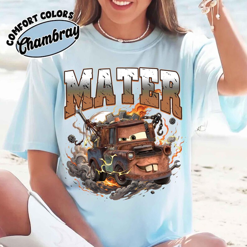 K&ouml;nnte beinhalten: Hellblaues T-Shirt mit einer Grafik von Hook aus dem Film Cars. Das Wort "MATER" ist im Used-Look &uuml;ber der Grafik aufgedruckt. Das Shirt tr&auml;gt auch die Worte "COMFORT COLORS Chambray" auf der oberen linken Seite.