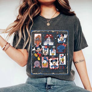 May include: A charcoal gray short-sleeve t-shirt featuring a graphic print of various cartoon characters in patriotic red, white, and blue outfits. The design includes multiple framed illustrations of characters, a castle, and the text 2026.