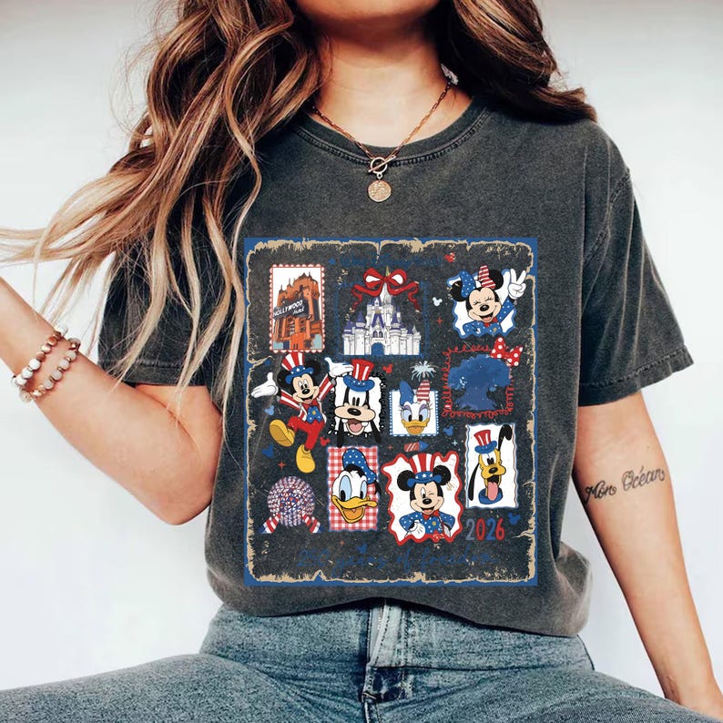May include: A charcoal gray short-sleeve t-shirt featuring a graphic print of various cartoon characters in patriotic red, white, and blue outfits. The design includes multiple framed illustrations of characters, a castle, and the text 2026.