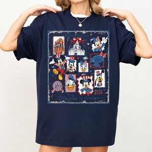 May include: A navy blue t-shirt featuring a graphic print of various cartoon characters in patriotic red, white, and blue outfits. The design includes a castle, fireworks, and the text 250 years of freedom and 2026, arranged in a collage style.