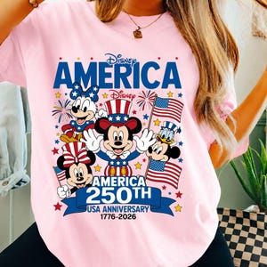May include: A light pink t-shirt featuring a graphic of Disney characters including Mickey Mouse, Minnie Mouse, and Donald Duck wearing patriotic red, white, and blue outfits. The text reads Disney AMERICA 250TH USA ANNIVERSARY 1776-2026.