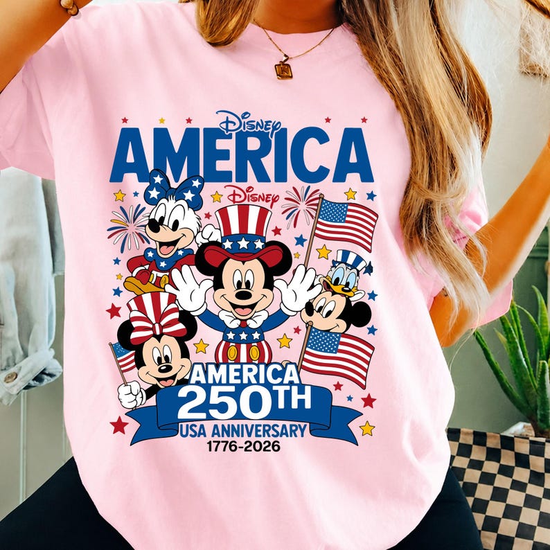 May include: A light pink t-shirt featuring a graphic of Disney characters including Mickey Mouse, Minnie Mouse, and Donald Duck wearing patriotic red, white, and blue outfits. The text reads Disney AMERICA 250TH USA ANNIVERSARY 1776-2026.