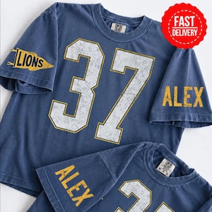 May include: A blue short-sleeved t-shirt featuring the number 37 in large white distressed font with gold outlines. The left sleeve displays a yellow pennant graphic with the word LIONS, while the right sleeve features the name ALEX in yellow.