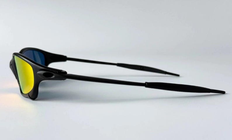 May include: Black sunglasses with a sleek, modern design. The lenses have a yellow-gold mirrored finish on one side and a blue tint on the other. The frames and arms are black, creating a bold contrast against the light background.