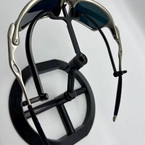 Puede incluir: Gafas de sol con montura plateada y lentes oscuras sobre un expositor negro. Las gafas de sol tienen un dise&ntilde;o elegante y moderno con patillas curvas y un puente. El expositor es de dise&ntilde;o minimalista.