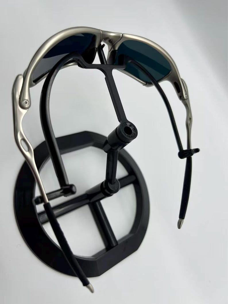 Puede incluir: Gafas de sol con montura plateada y lentes oscuras sobre un expositor negro. Las gafas de sol tienen un dise&ntilde;o elegante y moderno con patillas curvas y un puente. El expositor es de dise&ntilde;o minimalista.