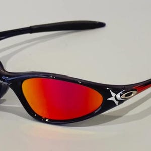 May include: Black and navy sunglasses with a star design and red and yellow lenses. The sunglasses have a curved frame and are designed for sports or outdoor activities. The lenses transition from yellow to red.