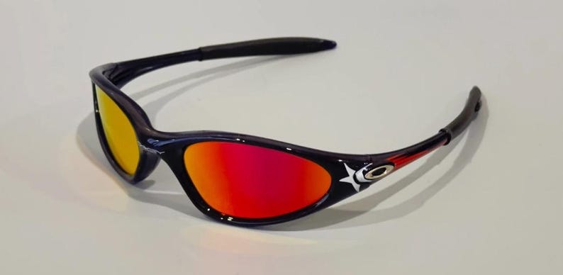 May include: Black and navy sunglasses with a star design and red and yellow lenses. The sunglasses have a curved frame and are designed for sports or outdoor activities. The lenses transition from yellow to red.