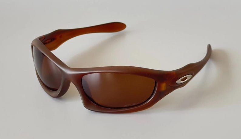 May include: Brown sunglasses with a curved frame and tinted lenses. The sunglasses have a sleek, sporty design, with a small silver emblem on the temple. The lenses and frame are a matching shade of brown.