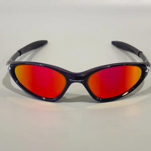 May include: Black sunglasses with a curved frame and vibrant red-orange lenses. The sunglasses have a star detail on the temples and the brand name "Oakley" is visible on the bridge.