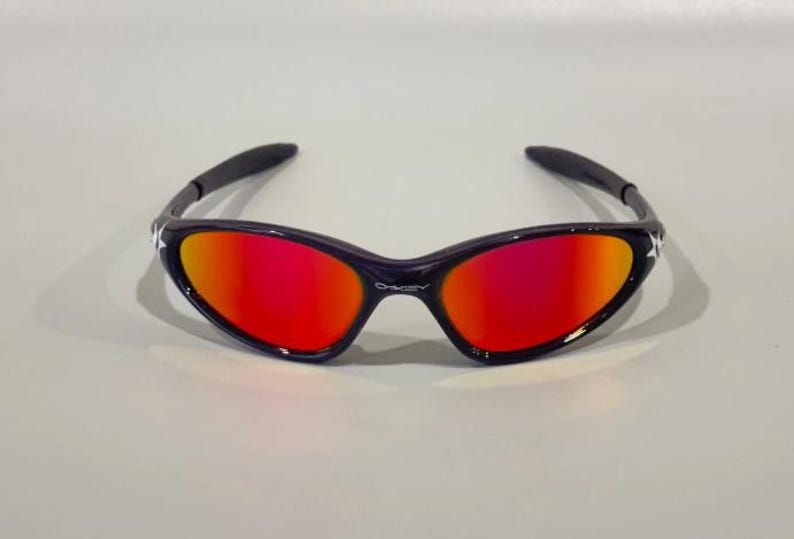 May include: Black sunglasses with a curved frame and vibrant red-orange lenses. The sunglasses have a star detail on the temples and the brand name "Oakley" is visible on the bridge.