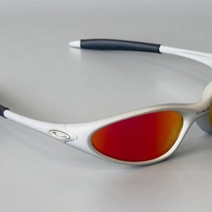 K&ouml;nnte beinhalten: Sonnenbrille mit silbernem Rahmen und roten und gelben Gl&auml;sern. Die Sonnenbrille hat wei&szlig;e B&uuml;gel mit schwarzen, gummierten Spitzen. Das Oakley-Logo ist auf dem Rahmen sichtbar. Modernes, sportliches Design.