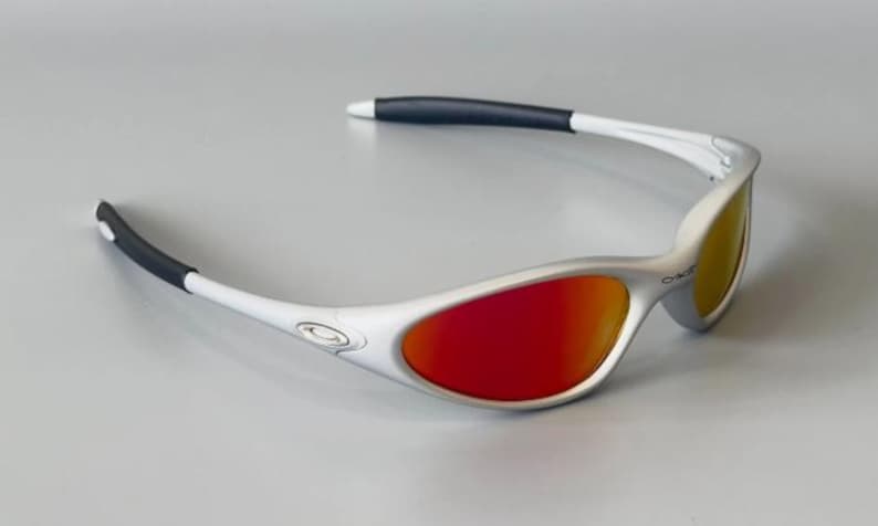 K&ouml;nnte beinhalten: Sonnenbrille mit silbernem Rahmen und roten und gelben Gl&auml;sern. Die Sonnenbrille hat wei&szlig;e B&uuml;gel mit schwarzen, gummierten Spitzen. Das Oakley-Logo ist auf dem Rahmen sichtbar. Modernes, sportliches Design.