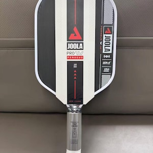 May include: A JOOLA Pro W Perseus pickleball paddle. The paddle face is black and white with vertical stripes and red accents. The handle is silver with the text "TAP HERE". The paddle is USA-A certified.