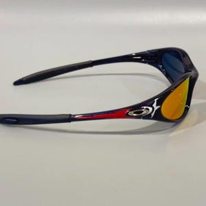May include: Sport sunglasses with a black frame, blue lenses, and orange-yellow mirrored lenses. The arms feature red and white accents with a logo. The sunglasses are designed for outdoor activities.
