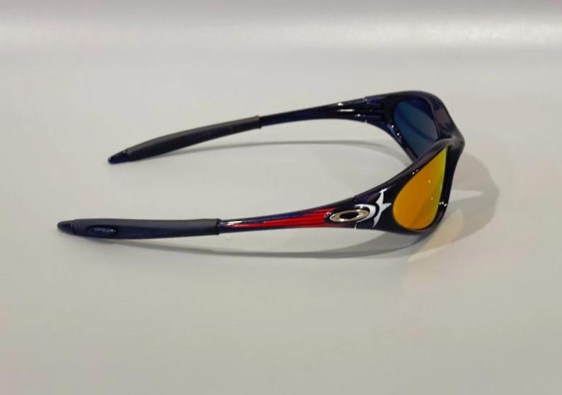 May include: Sport sunglasses with a black frame, blue lenses, and orange-yellow mirrored lenses. The arms feature red and white accents with a logo. The sunglasses are designed for outdoor activities.