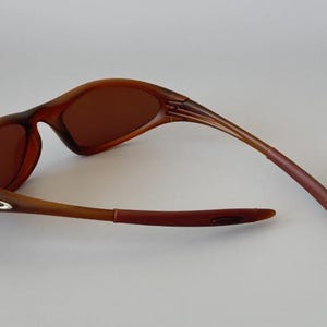 May include: Brown sunglasses with a curved frame and tinted lenses. The arms of the sunglasses are a darker shade of brown, and the lenses are a lighter shade of brown. The sunglasses are designed for outdoor use.