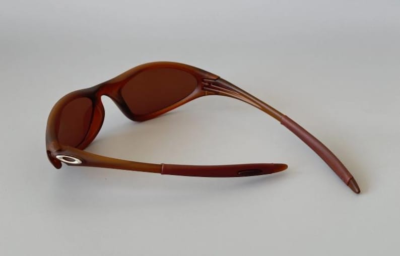 May include: Brown sunglasses with a curved frame and tinted lenses. The arms of the sunglasses are a darker shade of brown, and the lenses are a lighter shade of brown. The sunglasses are designed for outdoor use.