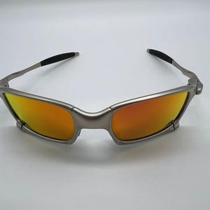 Puede incluir: Gafas de sol con montura plateada y lentes tintadas en naranja. Las gafas tienen un dise&ntilde;o moderno y angular con detalles negros en las patillas. Las lentes son de color naranja vibrante.